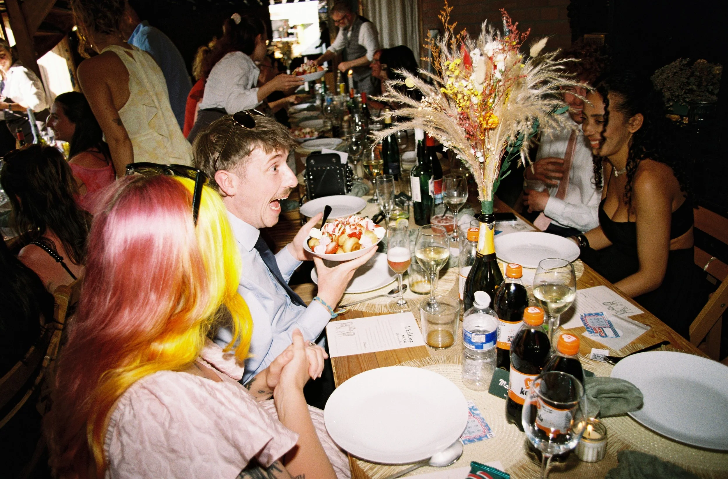 Candid wedding dinner moment captured by Elizabeth Royse Wedding Photography, showing guests laughing and chatting around a long rustic table filled with wine glasses, colourful florals and shared food, reflecting the joyful, relaxed atmosphere of a 