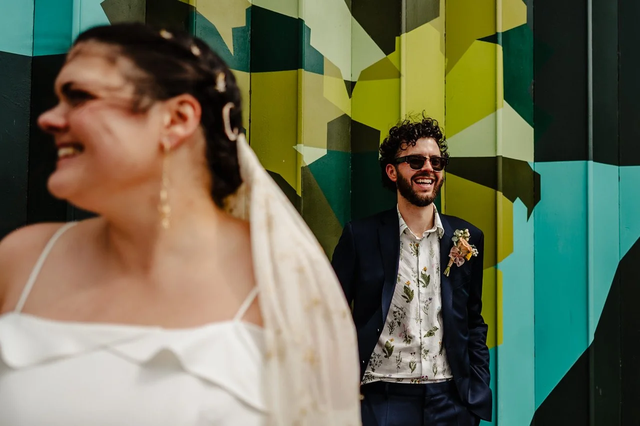 A joyful newlywed moment unfolds against a bold geometric mural where the groom leans back laughing in his floral shirt and navy suit while the bride turns toward him out of focus in the foreground her smile mirroring his and her veil catching the li