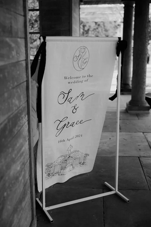 Monochrome fabric wedding banner by Blue Pumpkin Calligraphy, featuring elegant hand-lettered script and a bespoke illustration of the ceremony venue. The minimalist black-and-white palette and flowing calligraphy create a timeless and romantic welco