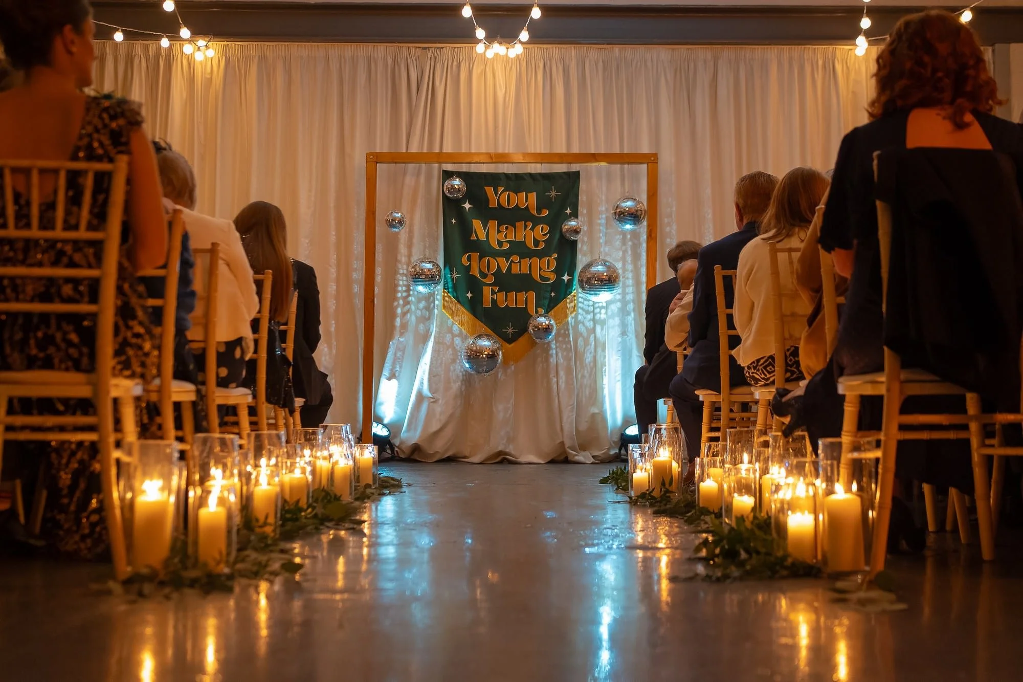 A softly lit aisle lined with glowing candles leads to a draped backdrop where a green and gold banner reading You Make Loving Fun hangs among gleaming disco balls, and guests seated on wooden chairs face forward in the warm ambience created by strin