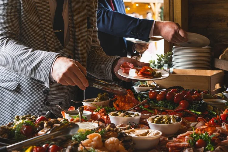 Guests serve themselves from a lavish buffet prepared by Regis Banqueting, featuring an assortment of Mediterranean-inspired dishes, fresh seafood, olives, roasted vegetables, and charcuterie. This vibrant wedding catering setup showcases the company