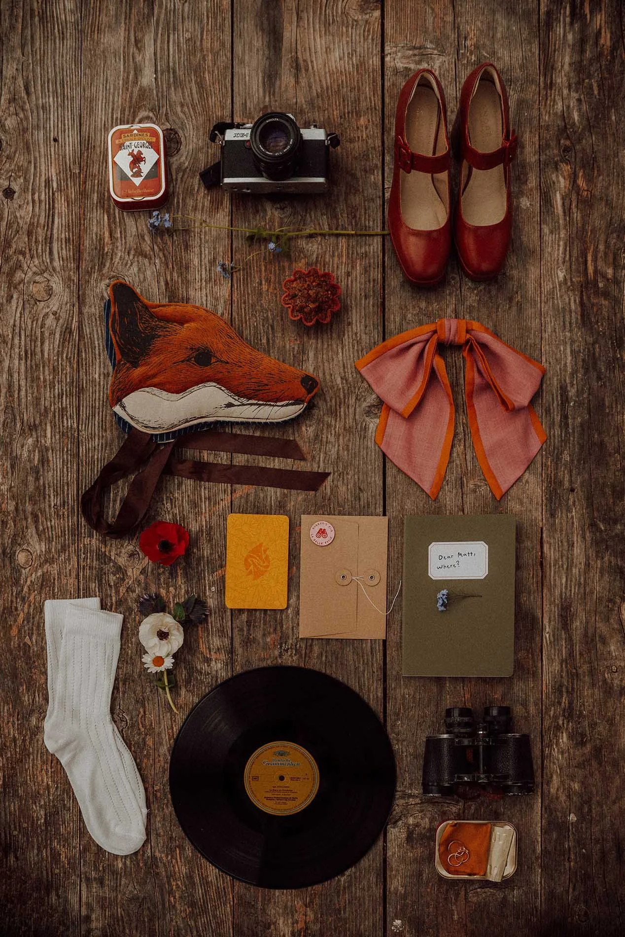 A characterful Wes Anderson inspired wedding flatlay featuring vintage props arranged on rustic wooden boards including a retro film camera, red Mary Jane shoes, a fox mask, patterned bow, vinyl record, binoculars, stationery, flowers and playful kee