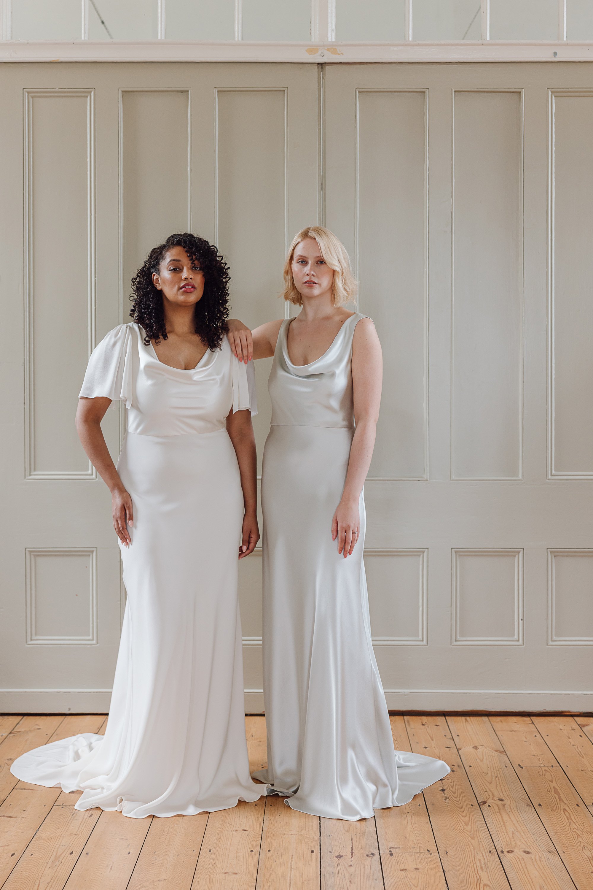 The two brides stand side by side in a softly lit room with pale panelled walls and warm wooden floors, each wearing a fluid silk gown that falls in clean, elegant lines, one with gentle draped sleeves and a relaxed cowl neckline and the other with a