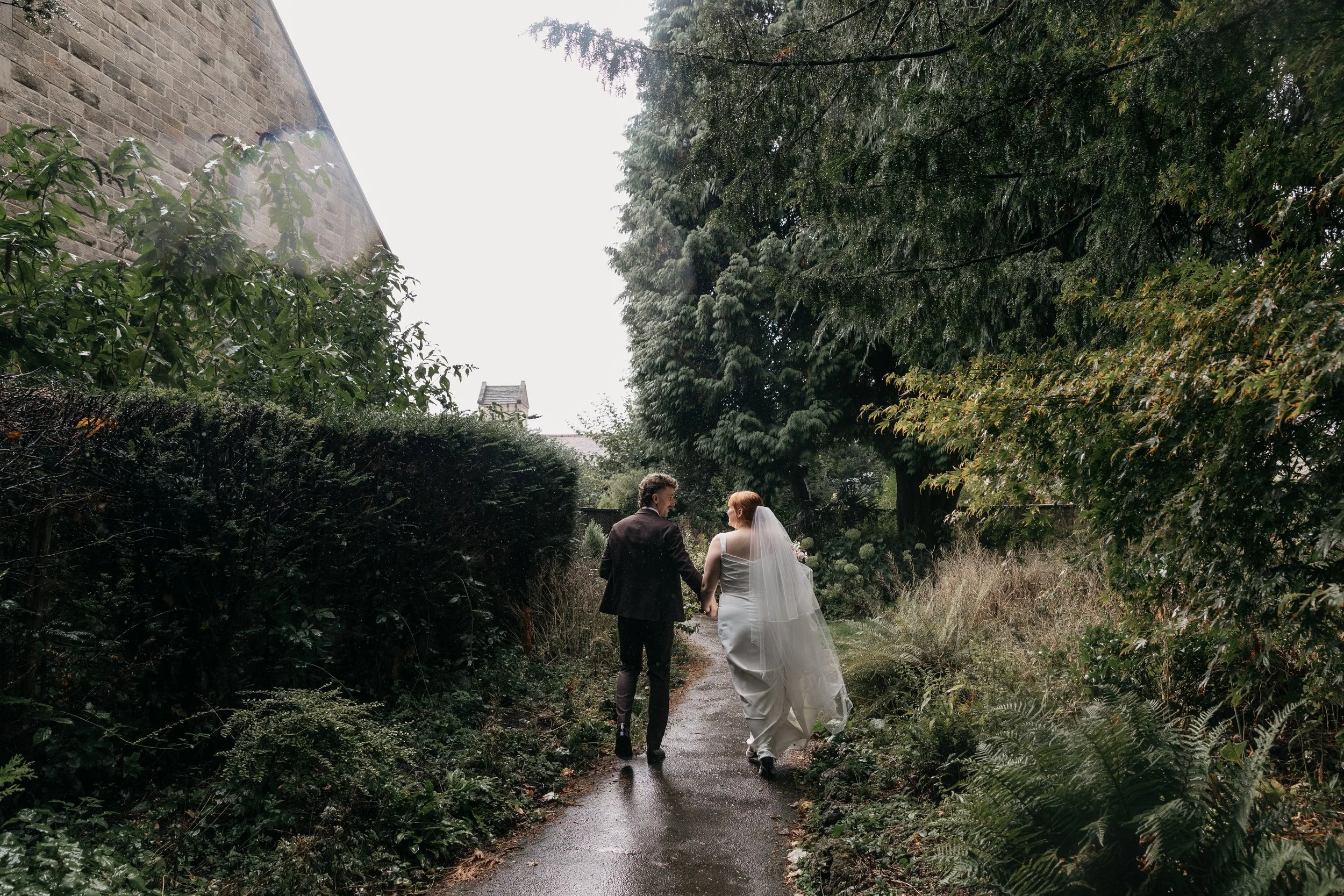 A tender and atmospheric moment captured by YU Photography, showing the couple walking hand in hand down a quiet, rain soaked garden path. The bride’s veil trails softly behind her while lush greenery arches overhead, creating a natural tunnel that f