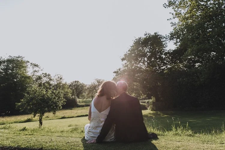 A peaceful and romantic sunset moment captured by YU Photography, showing the couple sitting close together on a grassy hillside as warm evening light glows behind them. With the bride leaning gently into her partner’s shoulder and the landscape stre