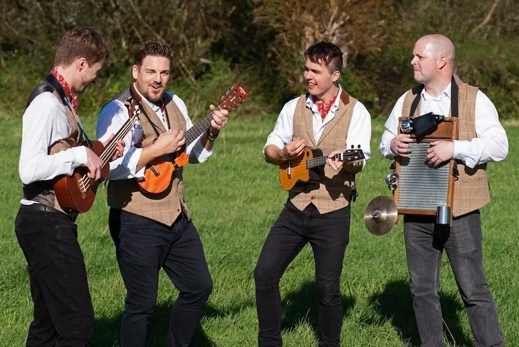 The Ukes of Hazard perform outdoors in a sunny field, bringing their trademark charm and energy to a live wedding performance. The four-piece band features ukulele players and a percussionist with a washboard drum, combining upbeat folk and acoustic 