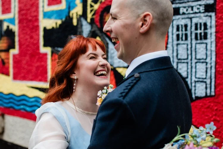 A colourful, joy-filled wedding portrait captured by Mirrorbox Photography, showing a couple laughing together in front of a bright, urban mural. The bride’s striking red hair and pastel dress contrast beautifully with the bold graffiti-style artwork