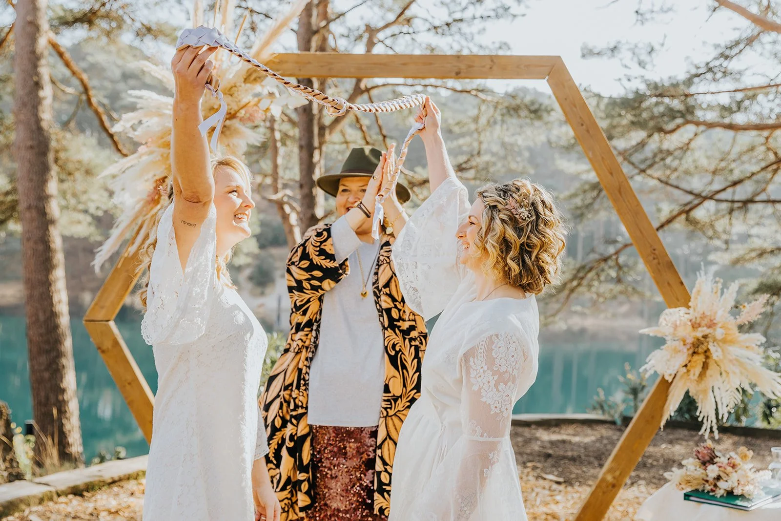Two happy brides with their celebrant in front of a hexagonal altar piece at their outdoor wedding, styled by Wild Soul Weddings, eco wedding planners in the UK. Lotus Photography UK 