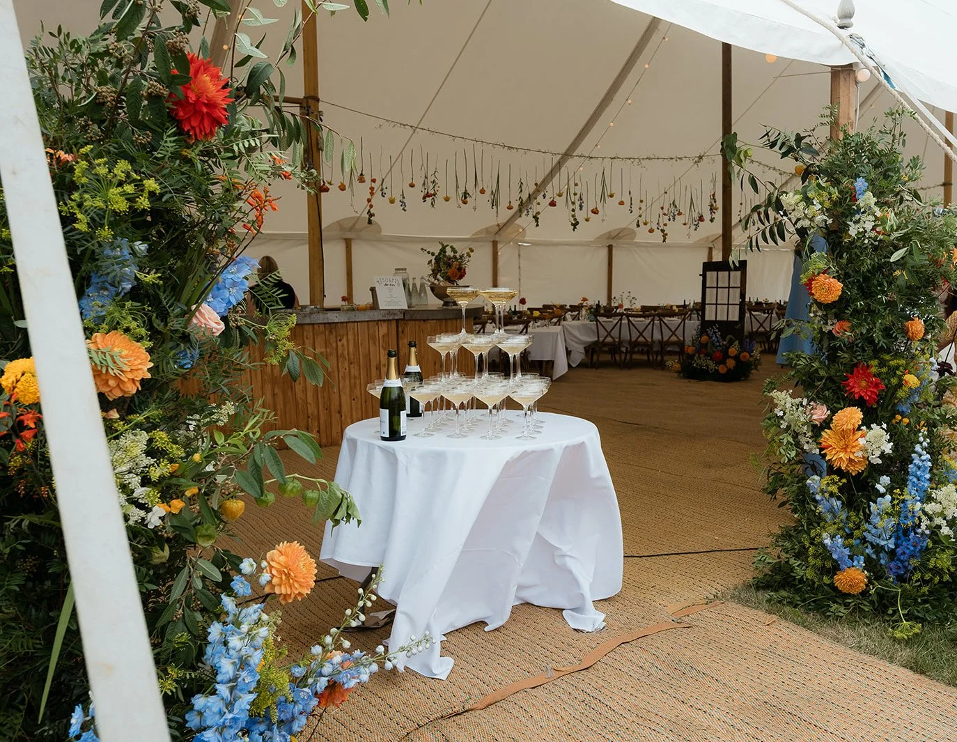 A table of champagne inside a tent, surrounded my colourful florals. Bella & George  - That Weddings Photo.