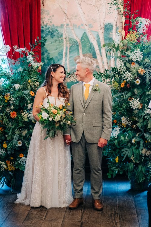 Colourful garden style wedding flowers by The Art Bunch featuring vibrant seasonal blooms, soft greenery and natural meadow inspired arrangements framing the couple during their ceremony. With the bride holding a textured bouquet in warm yellow and g