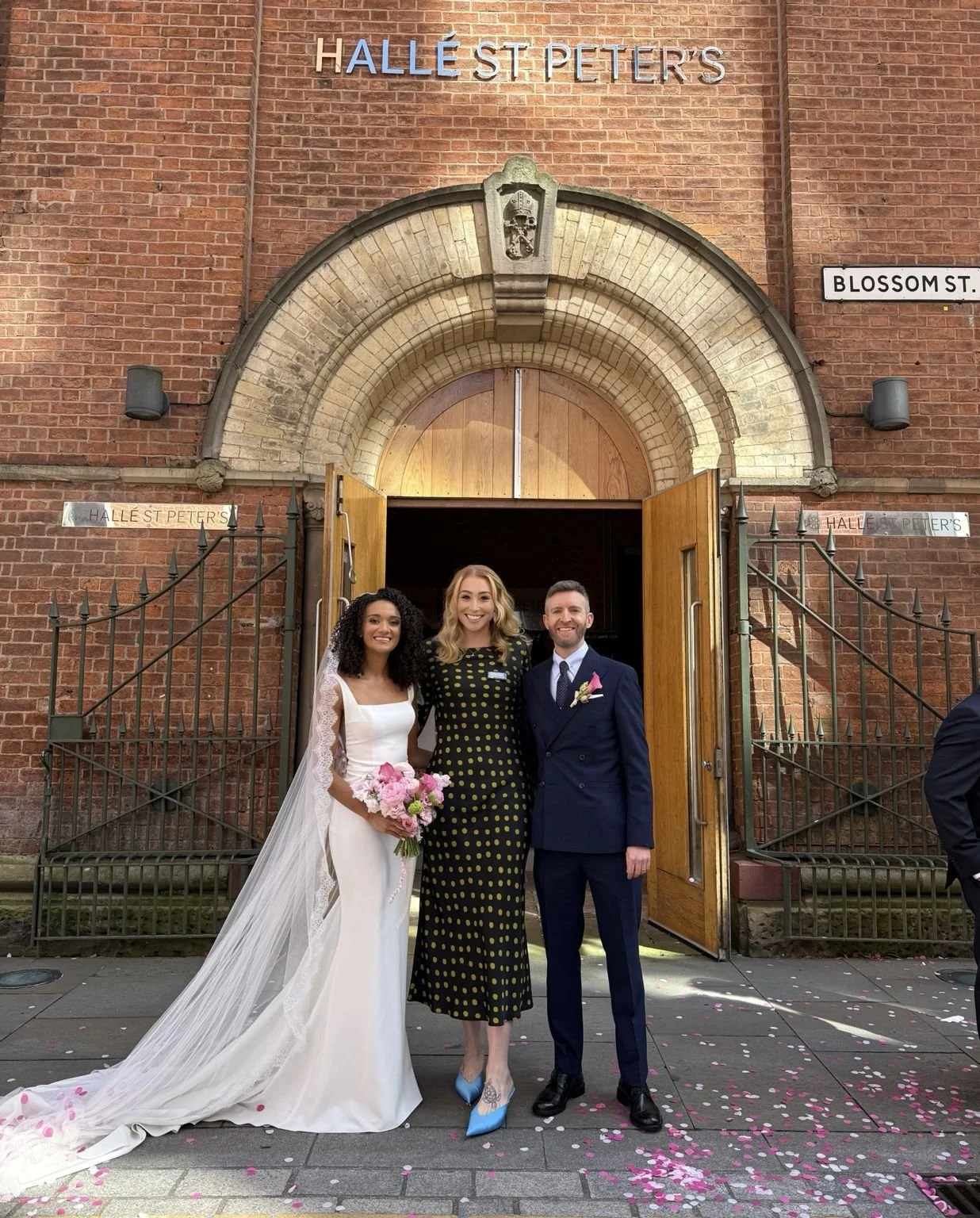 A photo of a celebrant in a black dress standing with her bride and groom outside Halle St Peters. Photo by Emma Crompton Weddings.