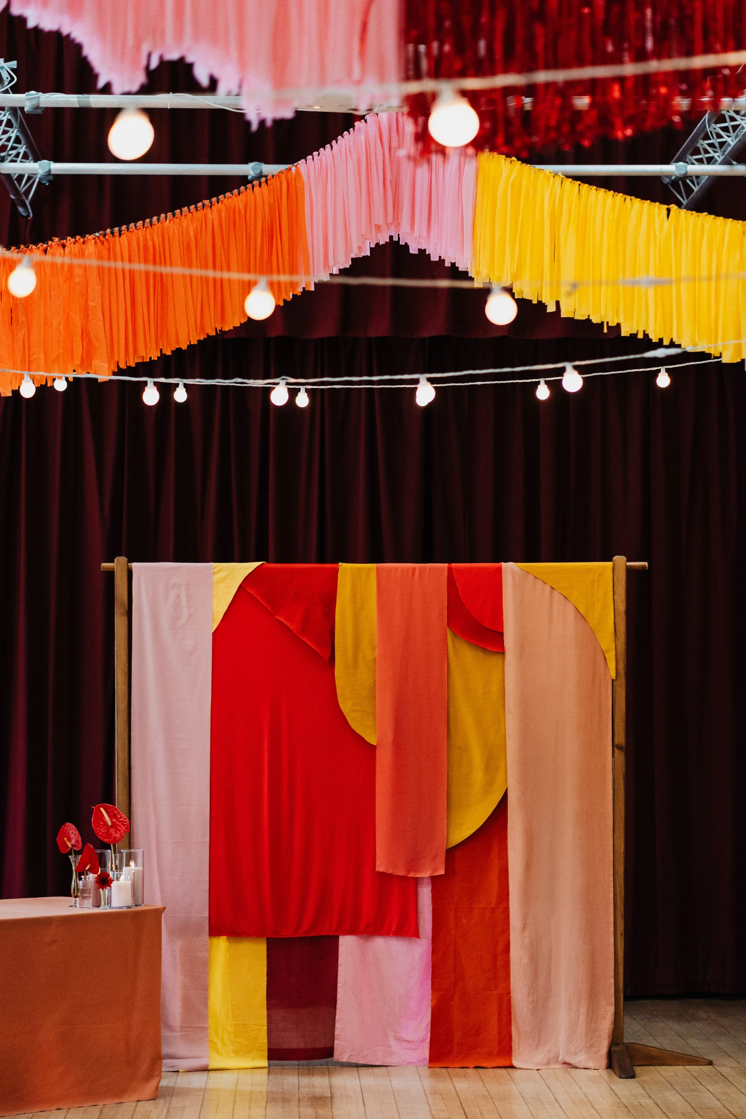 A bold, contemporary wedding backdrop featuring layered fabric panels in red, coral, peach, blush and yellow, draped asymmetrically over a freestanding frame. Above, rows of suspended fabric fringe in matching warm tones create a dramatic ceiling ins