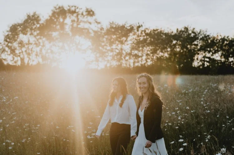 A warm and romantic outdoor wedding portrait captured by Matt Fox Photography, showing a couple walking hand in hand through a meadow at golden hour. The soft evening sunlight creates a beautiful glow around them, illuminating wildflowers and casting