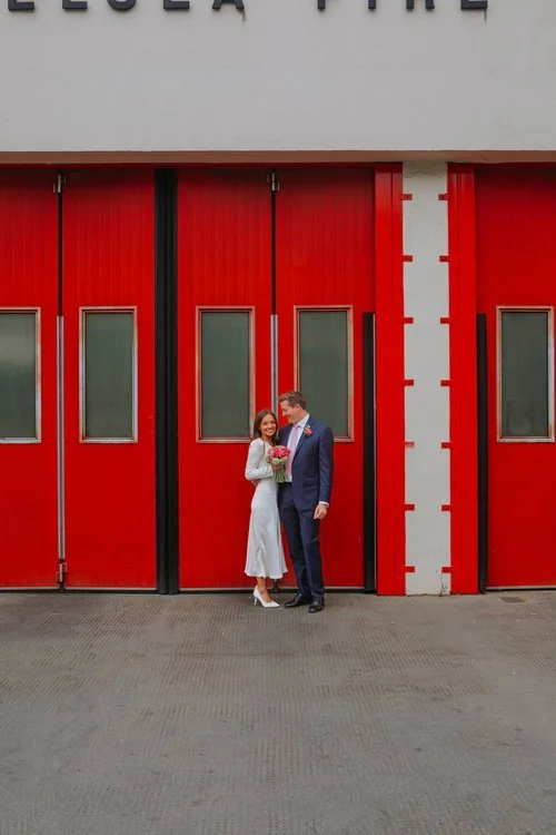 A couple stand smiling in front of bold red fire station doors, captured by Joshua Humphrey Photography. The striking urban backdrop and their elegant outfits create a stylish modern wedding portrait with a clean contemporary city feel.