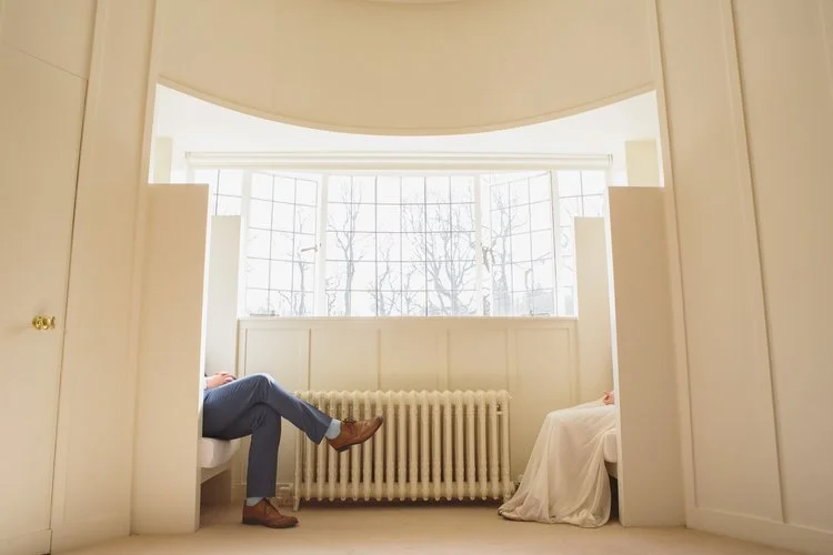 A modern, minimalist wedding photograph captured by Mirrorbox Photography, featuring a couple seated apart in a bright, architectural space filled with soft natural light. The image focuses on the clean lines of the room, the large bay window, and th