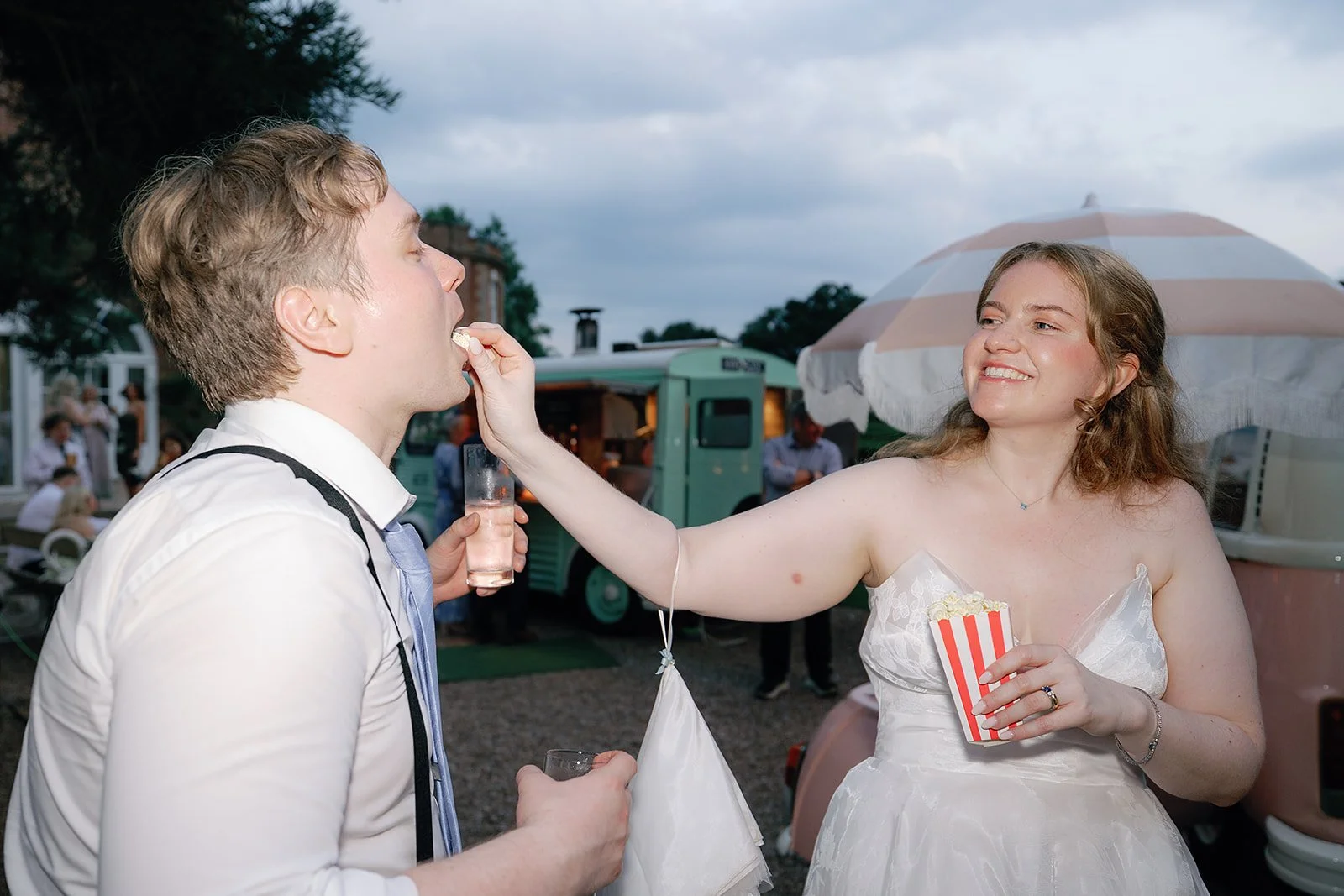 A playful candid moment crafted by Big Sister Events, featuring a bride and groom sharing popcorn and laughter against a charming outdoor setup with retro food trucks and pastel parasols creating a relaxed, joy filled vibe perfect for fun loving coup