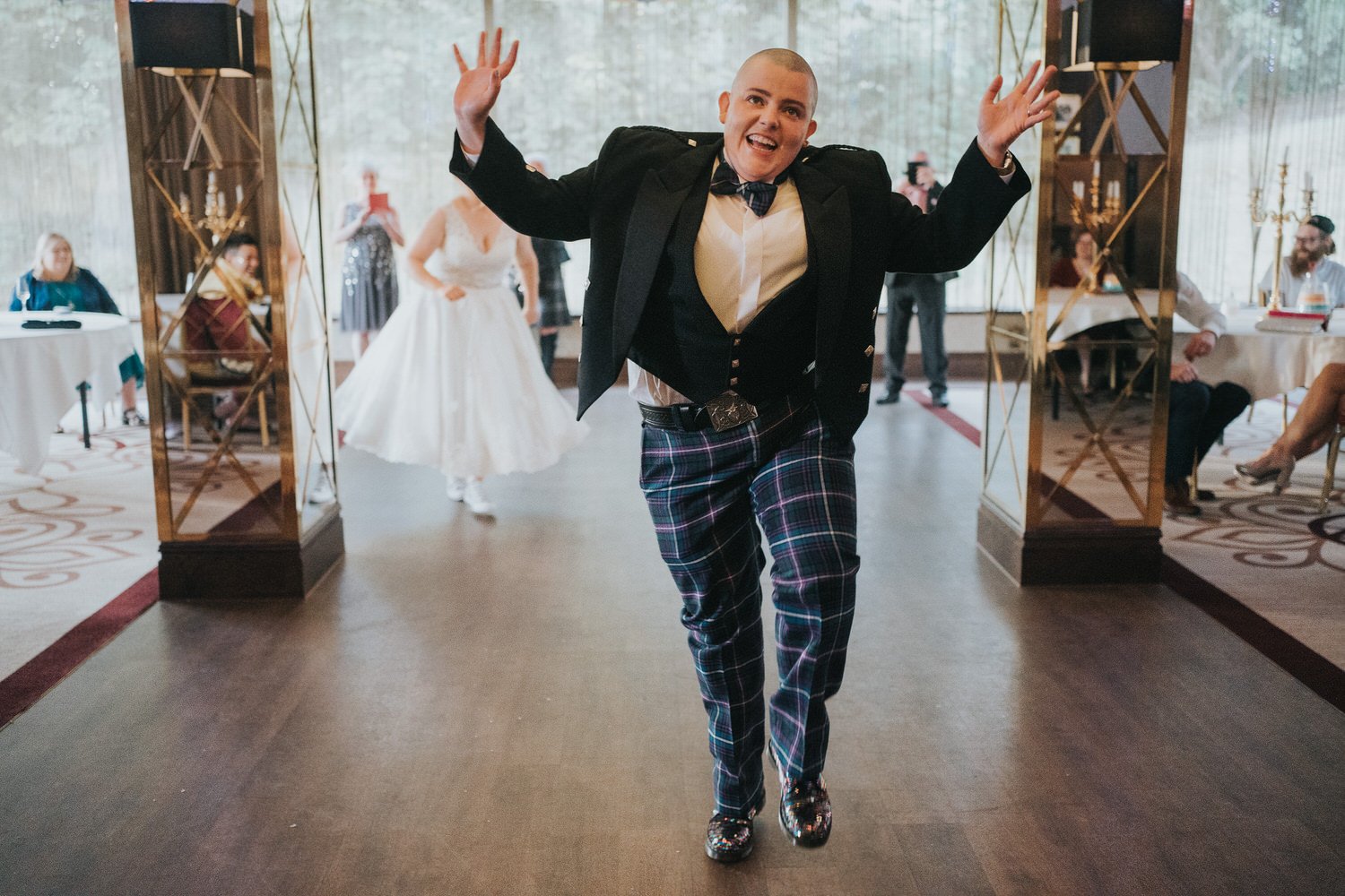 A high-energy wedding moment captured by Good Luck Wolf Photography, showing a guest in a tartan suit dancing exuberantly into the reception as the couple celebrate behind them, perfectly reflecting relaxed, documentary wedding photography full of pe