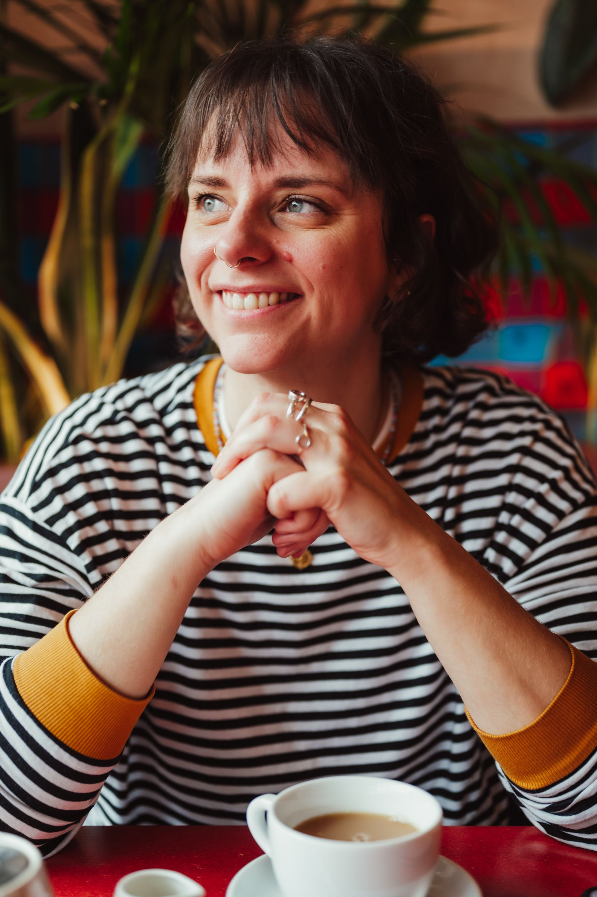 Cherry and Fern Celebrations smiles warmly while sitting in a relaxed café setting, enjoying a coffee. Her casual striped top and friendly expression capture her approachable and creative personality, ideal for couples seeking a down-to-earth and inc