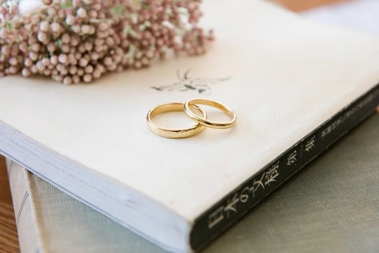 Handcrafted gold wedding bands by Nikki Stark Jewellery, displayed on a vintage book with delicate floral accents. Ethically made in the UK, these classic rings are perfect for modern couples seeking elegant, bespoke wedding jewellery.