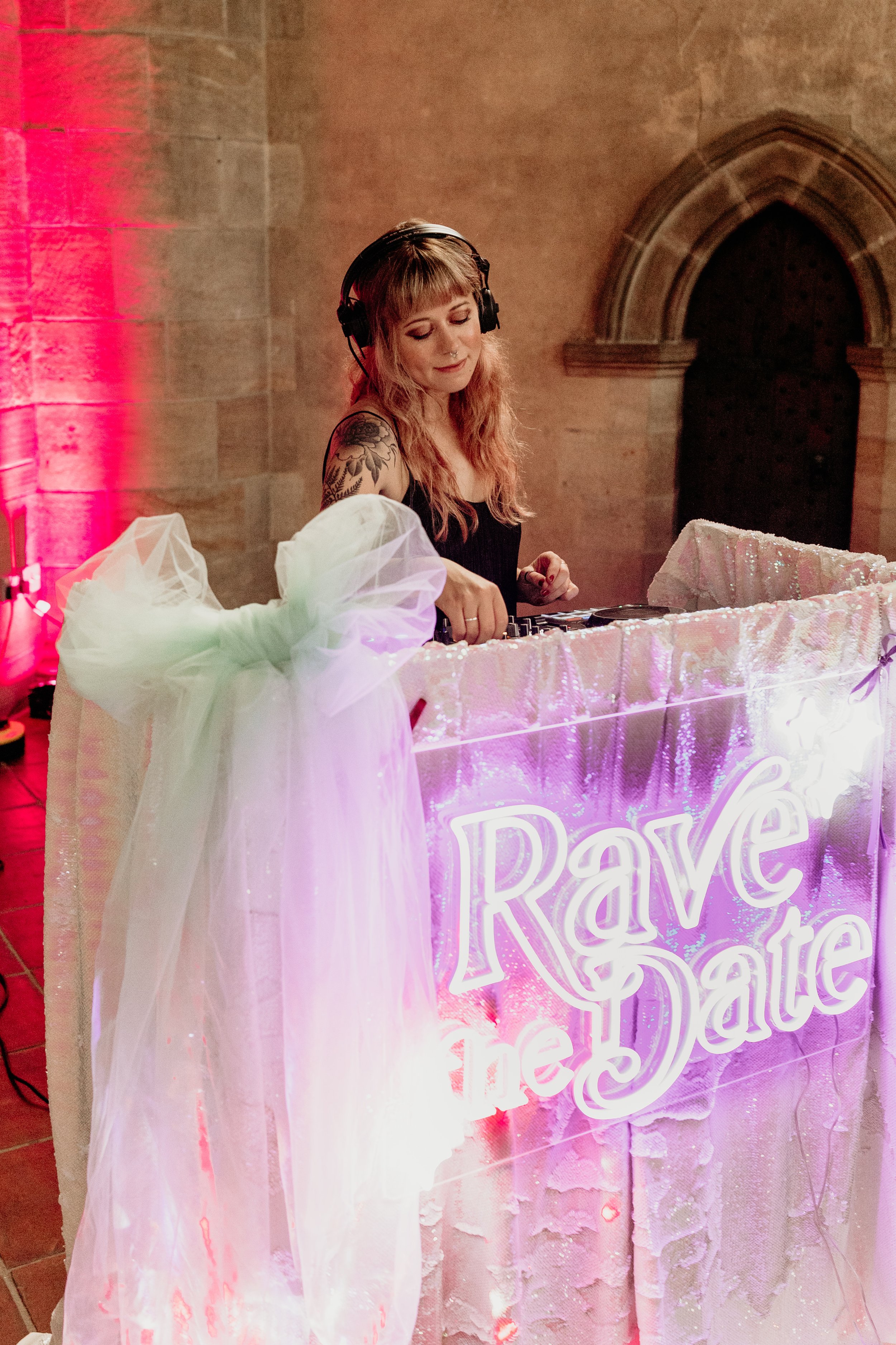 A stylish female wedding DJ from Rave the Date performing at an alternative wedding reception. She stands behind a shimmering sequin DJ booth decorated with soft tulle and a glowing neon sign that reads “Rave the Date.” The warm, atmospheric lighting