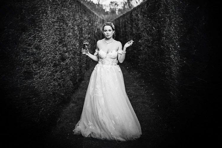 A gritty, high-contrast black and white photograph in Matt Parry Photography’s signature unapologetic documentary style. A bride stands centered between two towering hedges that form a narrow, dramatic corridor leading into the distance. She holds a 