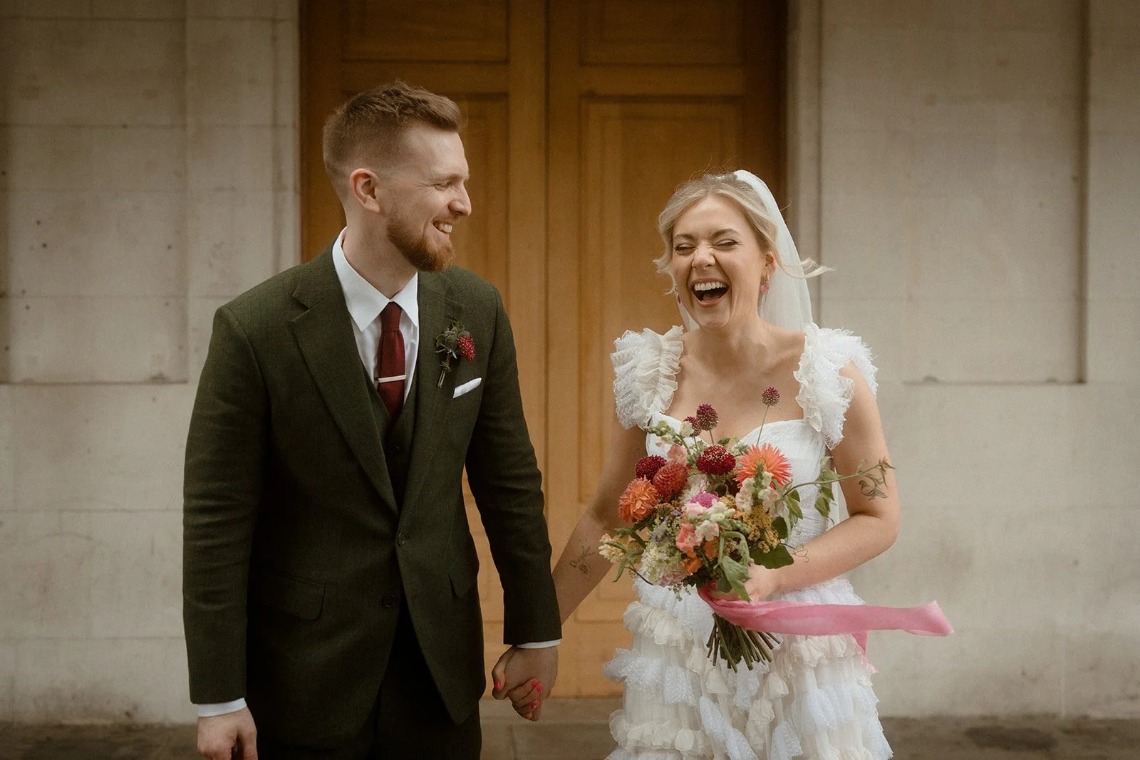 A bride and groom stand hand in hand outside large wooden venue doors, with the groom in a green suit and the bride in a textured white dress holding a bouquet of bright flowers as both laugh and look at each other.