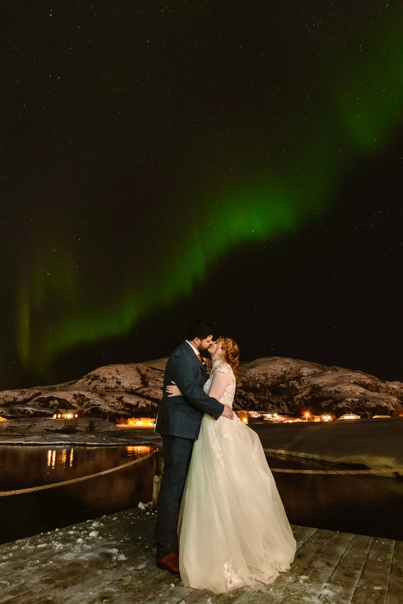 A romantic winter wedding photo captured by Bluebell Photo Studio, featuring a bride and groom sharing a kiss beneath the glowing green Northern Lights in Norway. Set against snow-covered mountains and a star-filled sky, this magical Arctic wedding m