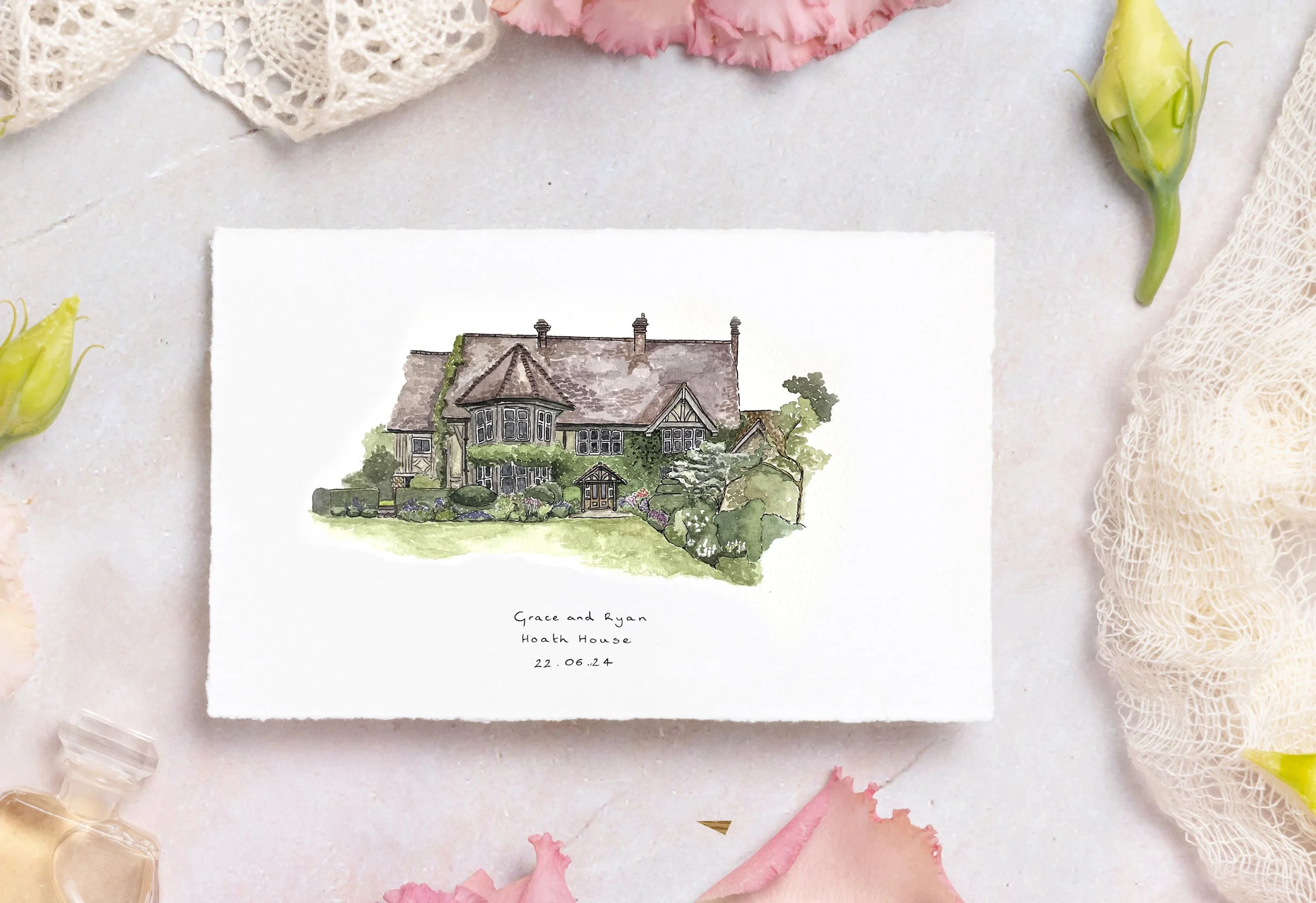 A delicate venue illustration card by Feather & Ink Stationery featuring a hand painted watercolour of Hoath House on textured deckle edged paper styled with soft lace, pastel flower petals and romantic props creating an elegant fine art aesthetic pe