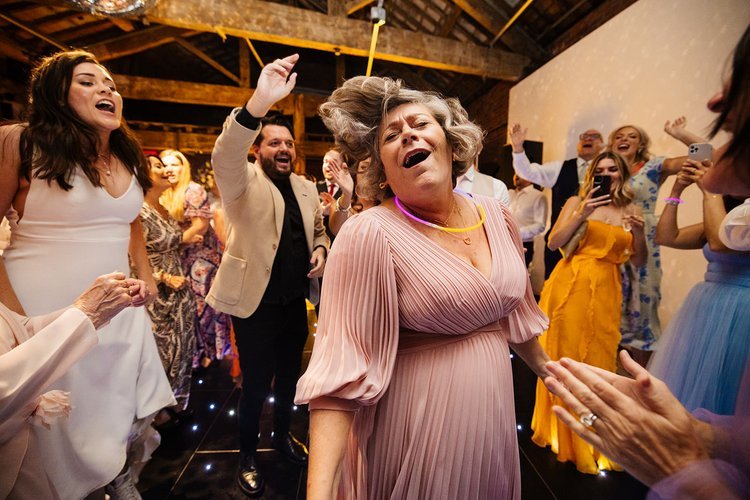 A lively wedding reception dance floor moment captured by Rachel Burt Photography, featuring guests in colourful outfits celebrating with full energy. A woman in a flowing pink dress dances at the centre with her hair flying, surrounded by cheering f