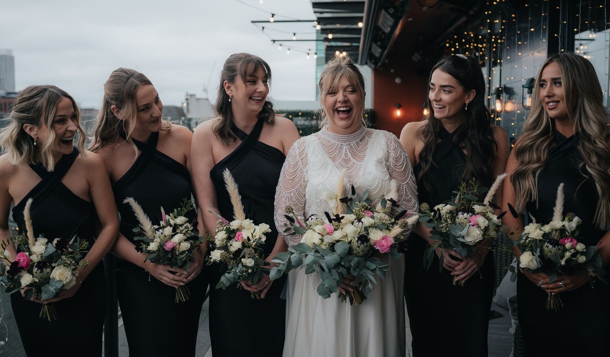 A beautiful bridal party wearing Rewritten Bridal Wear’s Roma dresses in classic black satin. The halter neckline with elegant cross-over detail gives each bridesmaid a sleek, contemporary silhouette that complements the bride’s vintage-inspired gown