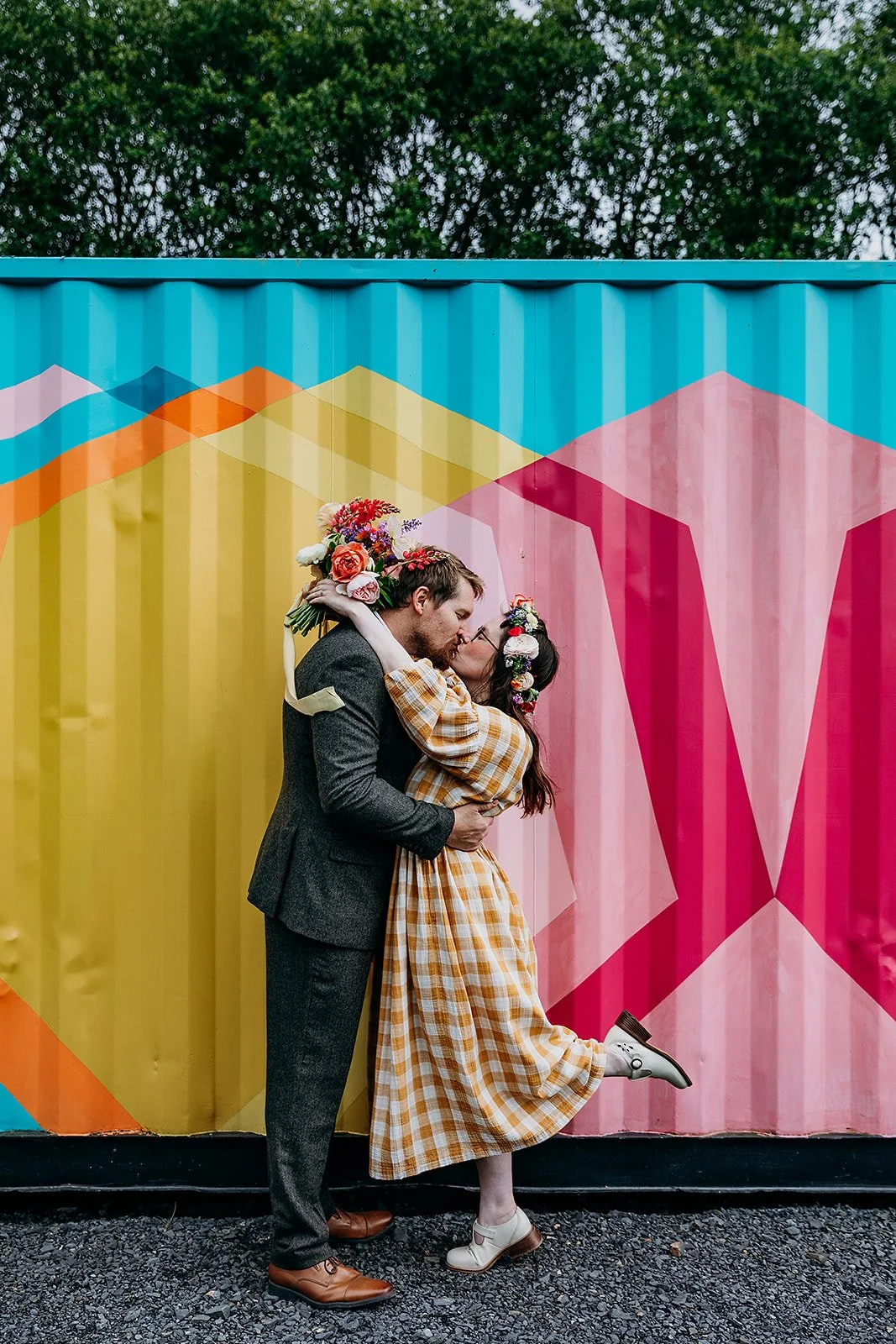 A colourful and joyful wedding portrait at The Giraffe Shed in Wales, featuring a newlywed couple sharing a kiss in front of the venue’s iconic geometric mural. The bride wears a yellow gingham dress with a floral crown and bouquet of bright blooms, 