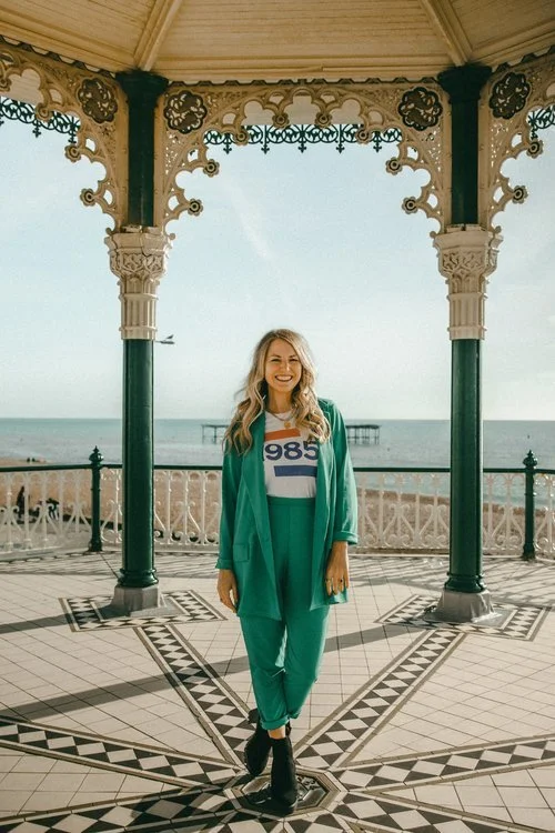 A smiling Roxy Celebrates Love stands under the ornate Brighton Bandstand with the sea and blue sky in the background. Dressed in a stylish teal suit and graphic T-shirt, she embodies a relaxed, modern approach to wedding ceremonies. This image captu