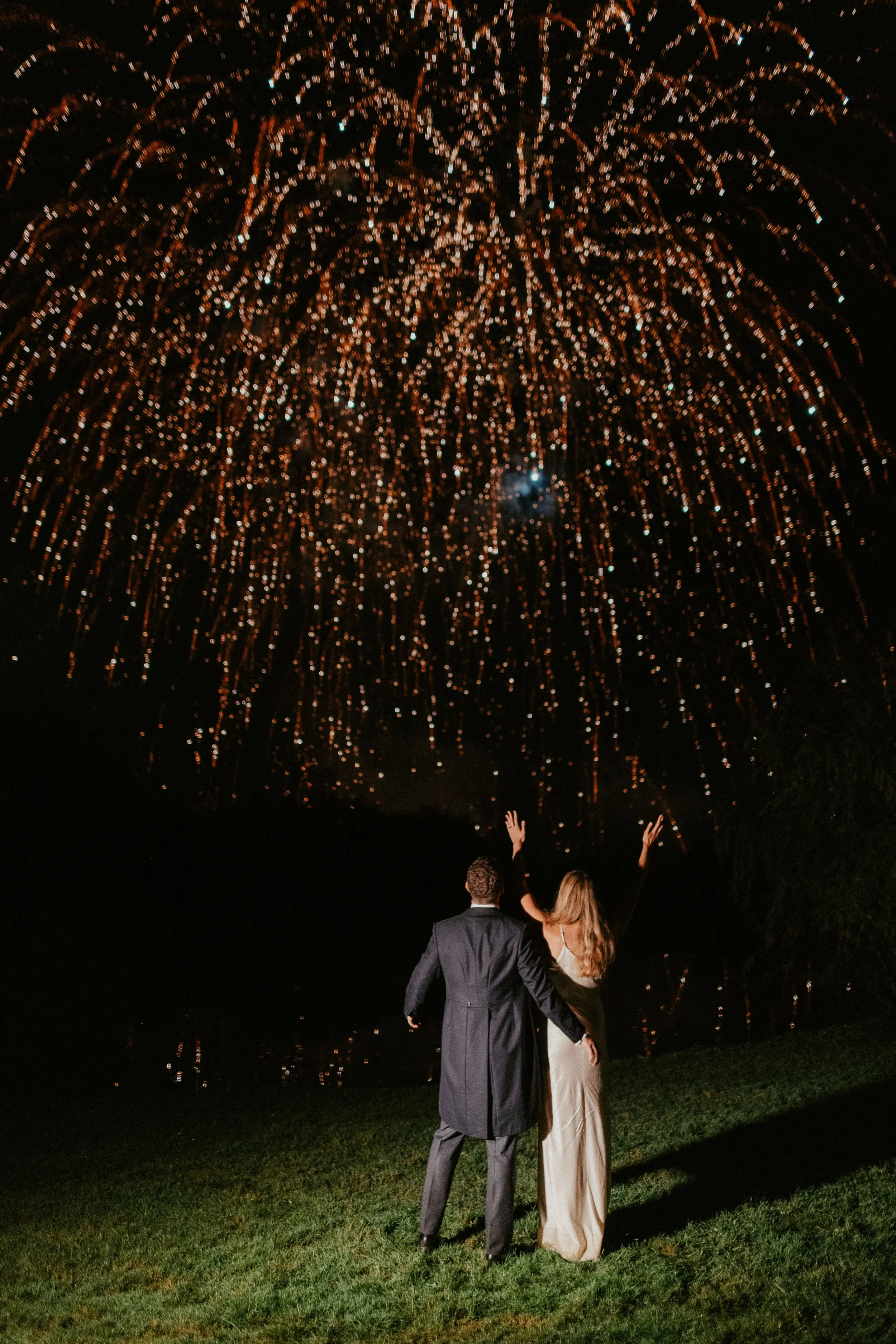 A newlywed couple stand arm-in-arm in front of a breathtaking fireworks display, the night sky exploding into cascades of golden light above them. The bride, in a sleek ivory gown, throws her hands joyfully into the air while her partner pulls her cl