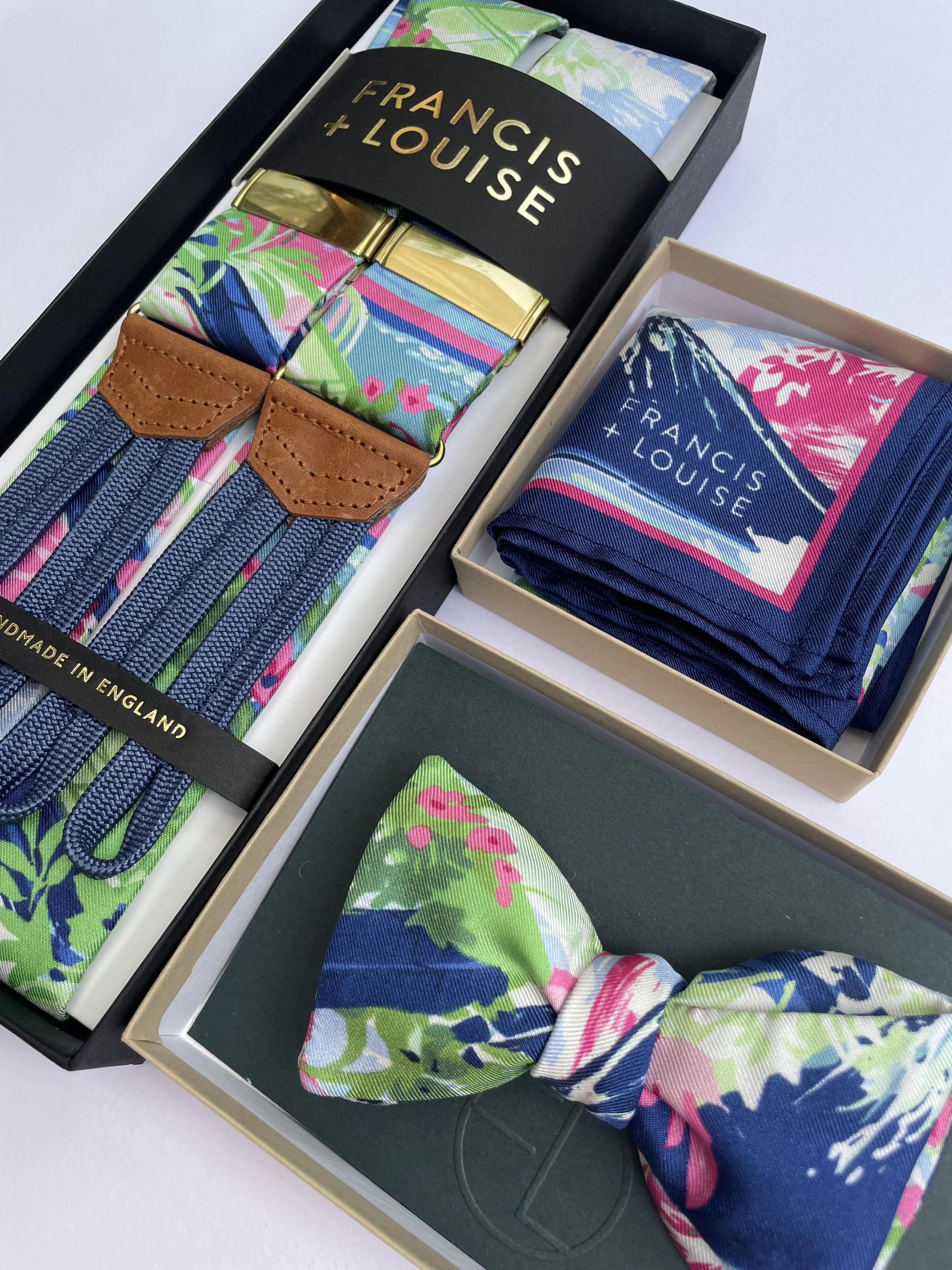 A coordinated Francis + Louise accessory set arranged in boxes, including patterned suspenders with gold clips and tan leather detailing, a folded pocket square featuring a blue landscape motif with pink and green accents, and a matching bow tie in v