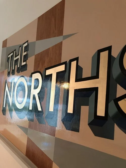 A close-up of a bespoke hand-painted wooden sign by Esther of Sylvie Signs featuring bold lettering that reads “THE NORTHS.” The design showcases a geometric background in warm tan, cream, and blue tones, with 3D shadowed typography that gives depth 