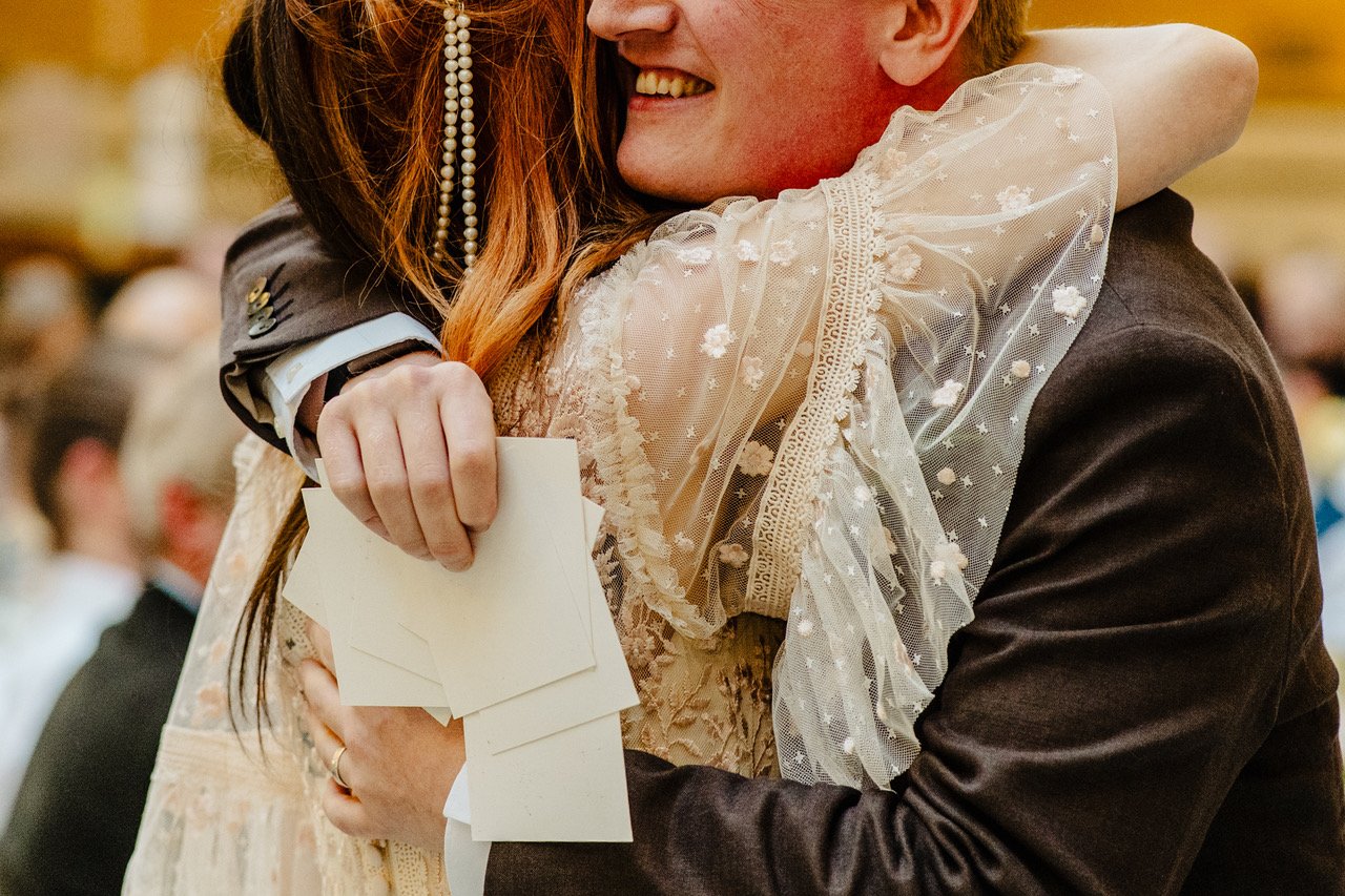 The photo captures an emotional embrace between two people as one wraps their arms tightly around the other clutching a small bundle of cards while delicate pearl strands and embroidered sheer sleeves frame the moment creating an intimate close up fi
