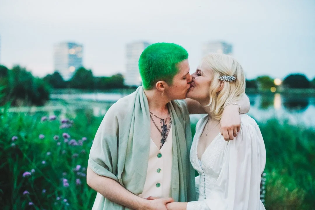 A tender and expressive outdoor wedding portrait captured by Barbara K Photography, showing a couple sharing a kiss beside a lakeside at twilight. One partner wears a soft, flowing outfit with a sage green shawl and vibrant green hair, while the othe