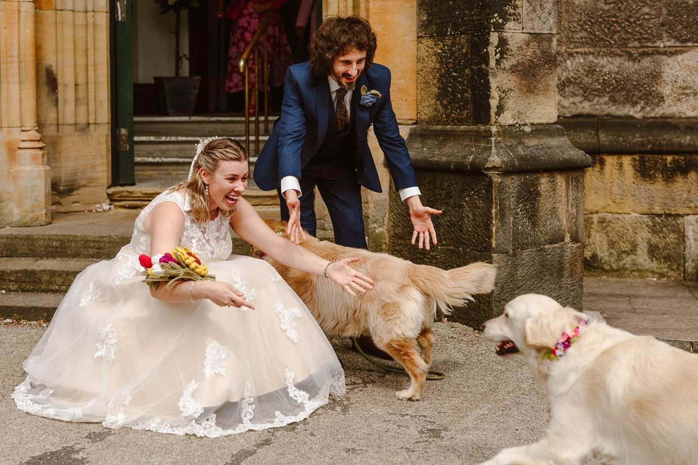 Fun candid wedding photo of a bride and groom greeting their excited dogs outside a historic UK church. The bride in a lace gown crouches down laughing as two golden retrievers run toward her, creating a joyful, natural moment perfect for couples loo