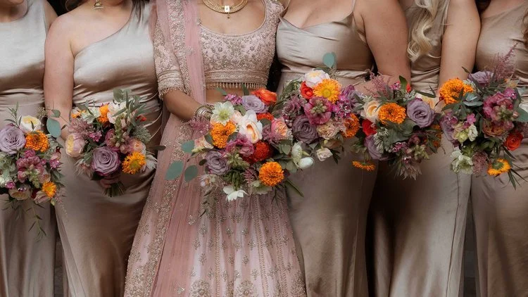 Elegant bridal party holding colourful wedding bouquets by New Bud Floristry featuring vibrant dahlias, roses, lisianthus and textured greenery in rich autumnal tones. Paired with champagne satin dresses and intricate bridal embroidery, these artisti