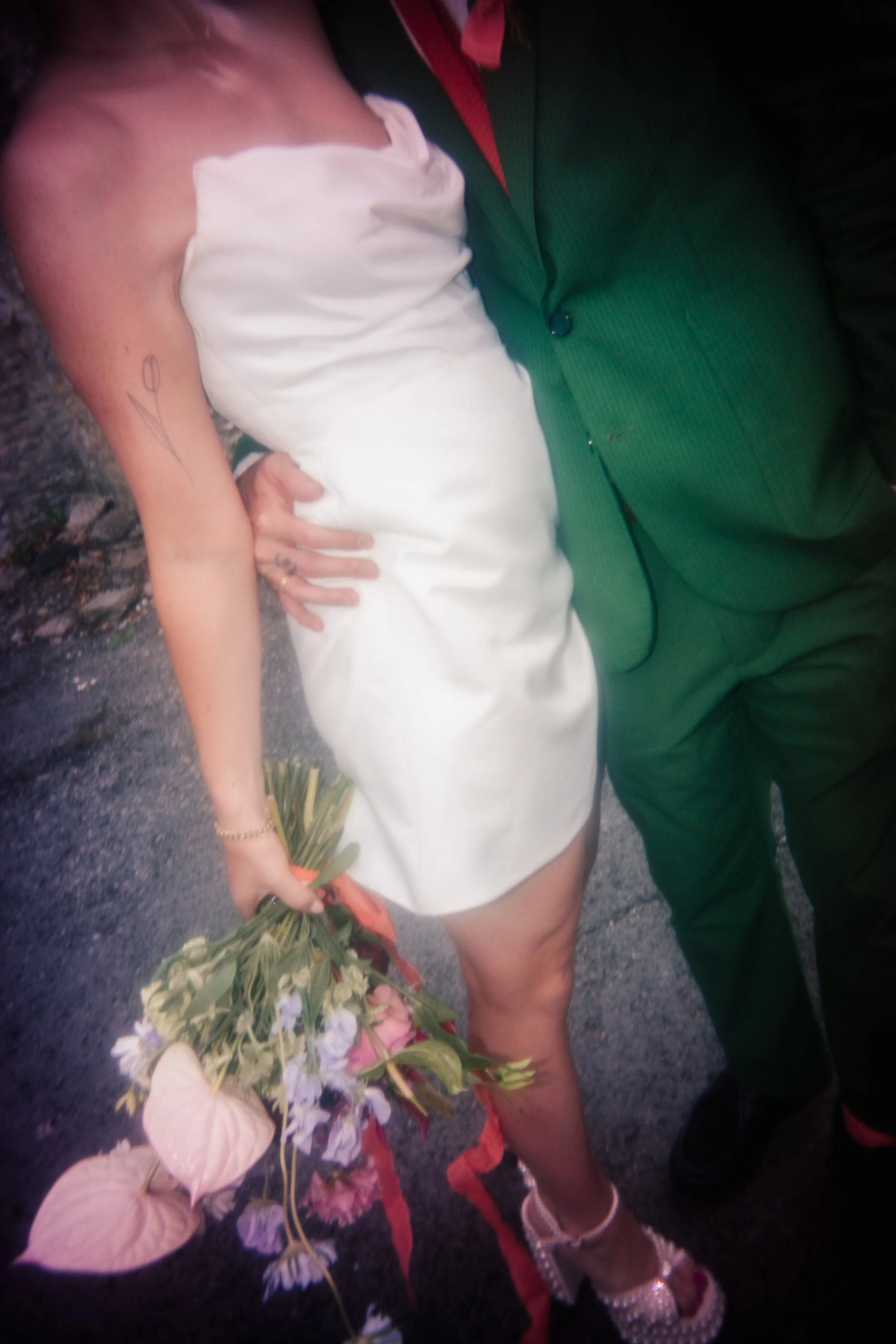 A creative disposable-camera style wedding photo showing a close-up of a bride in a short white satin dress holding a pastel bouquet with pink anthuriums and wildflowers, standing beside her partner in a green suit. The soft, dreamy blur adds a nosta