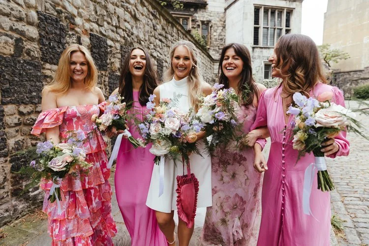 Joyful bridal party holding pastel seasonal bouquets by Botanique Workshop featuring soft roses, lilac scabiosa, peach dahlias and natural garden foliage. With the bride and bridesmaids walking together in vibrant pink dresses against historic stone 