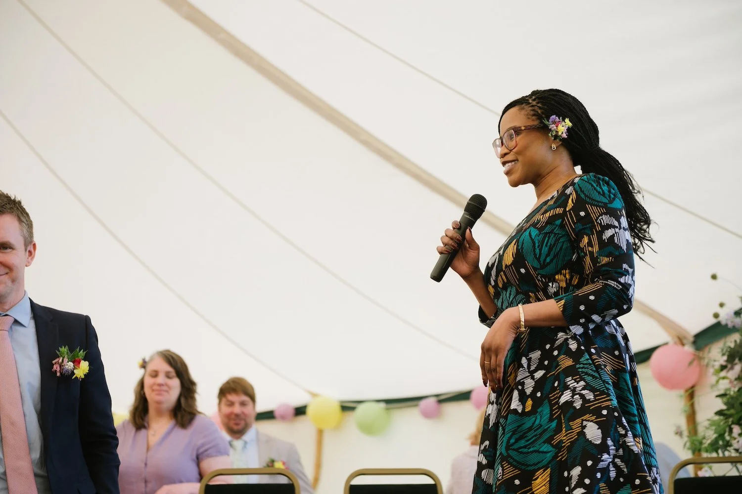 A Black woman called Deyi is smiling and holding a microphone. She is conducting a wedding ceremony. She wears glasses and has flowers in her hair. Photo by Mustard Yellow Photography.