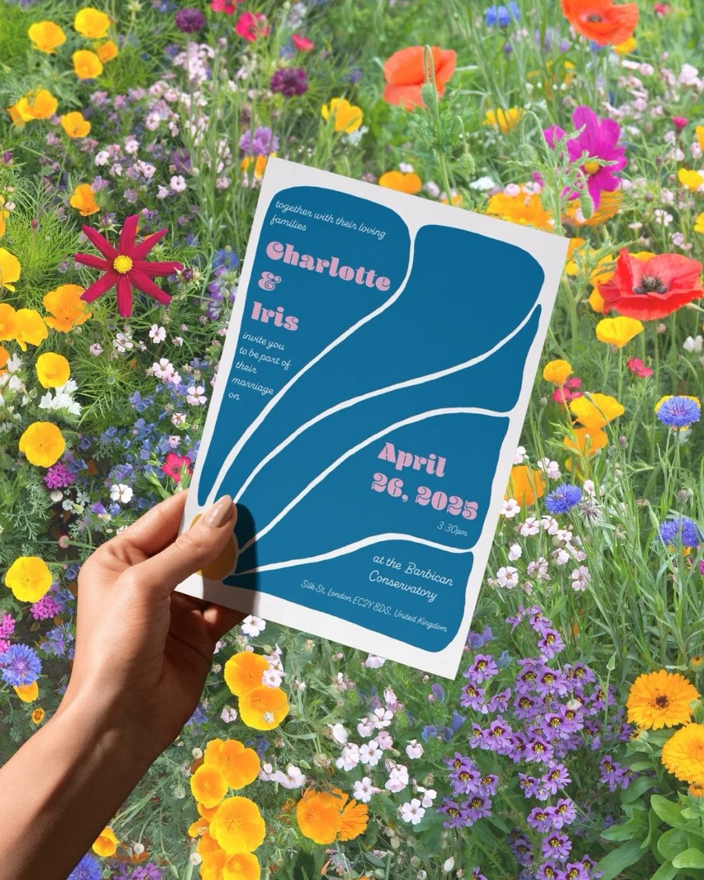A simple wedding invitation for 2 brides called Charlotte & Iris. A hand is holding it in a field of flowers.