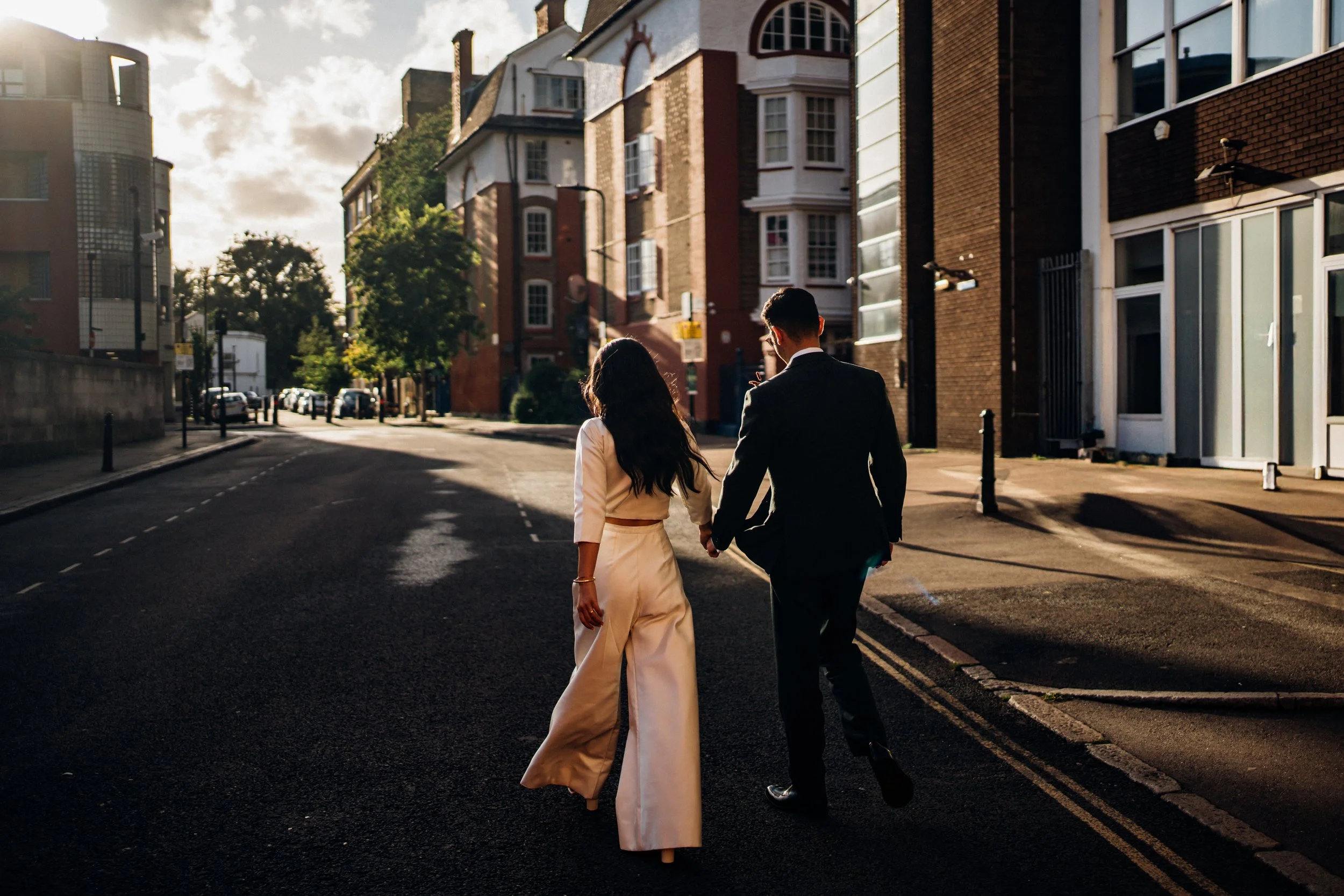 A bride and groom from behind. They are walking down the street and the bride is wearing a sating jumpsuit.