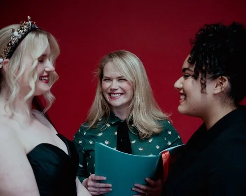 Wedding celebrant Una smiling warmly as she officiates a ceremony between two brides against a bold red backdrop. One bride wears a black gown with a jeweled crown, while the other wears a sleek black suit. A joyful and modern same-sex wedding celebr