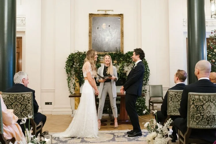A joyful couple exchange vows in an elegant indoor wedding ceremony, led by smiling celebrant Roxy, dressed in a stylish grey suit. The ceremony space features classic decor with lush green foliage and soft white flowers, creating a romantic and time