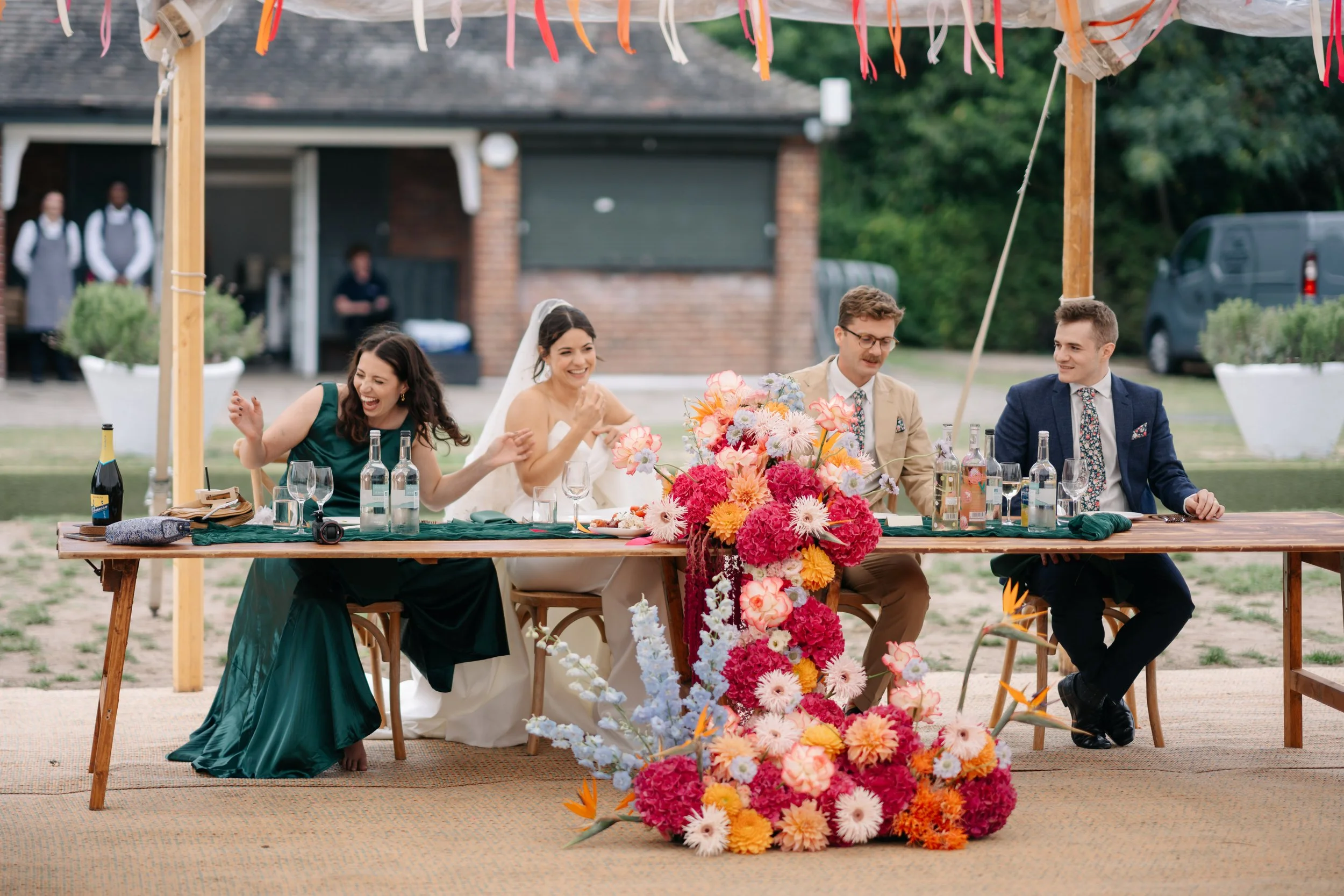 Joyful wedding head table captured by Tom Durn Photography, featuring a vibrant cascading floral arrangement by Sweet Pea Sorbet Floristry. The design includes pink, orange, and cream blooms, creating a bold and cheerful centrepiece for an outdoor ce