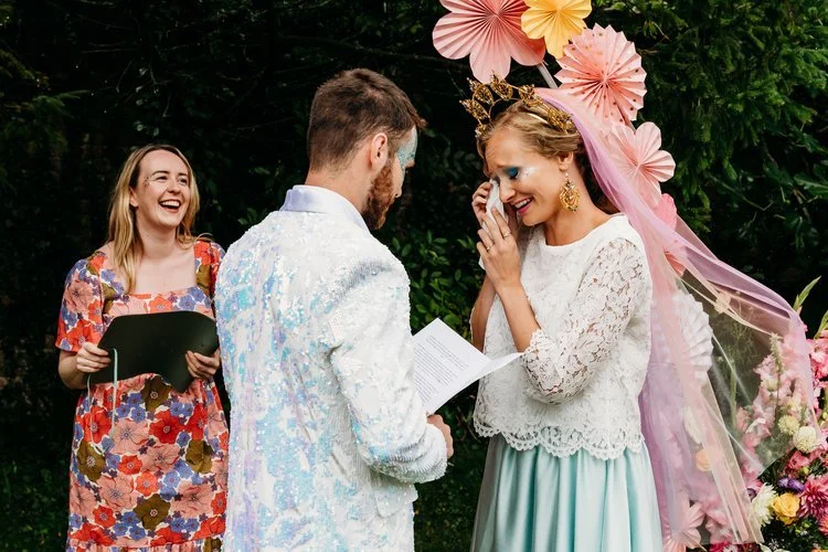 UK wedding celebrant Frances Celebrates You in a floral dress leads an emotional outdoor ceremony as a couple exchange vows beneath a colourful backdrop of paper flowers. The bride wipes away tears while the groom reads his vows, both dressed in crea
