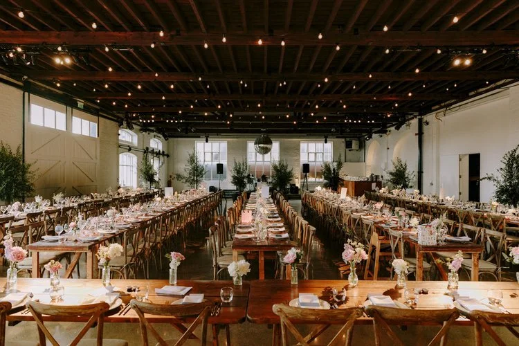 A beautifully styled wedding reception setup by Tasting Plates Wedding Caterers, featuring long rustic wooden tables adorned with elegant floral arrangements and soft lighting. The spacious venue is ready for guests to enjoy a refined dining experien