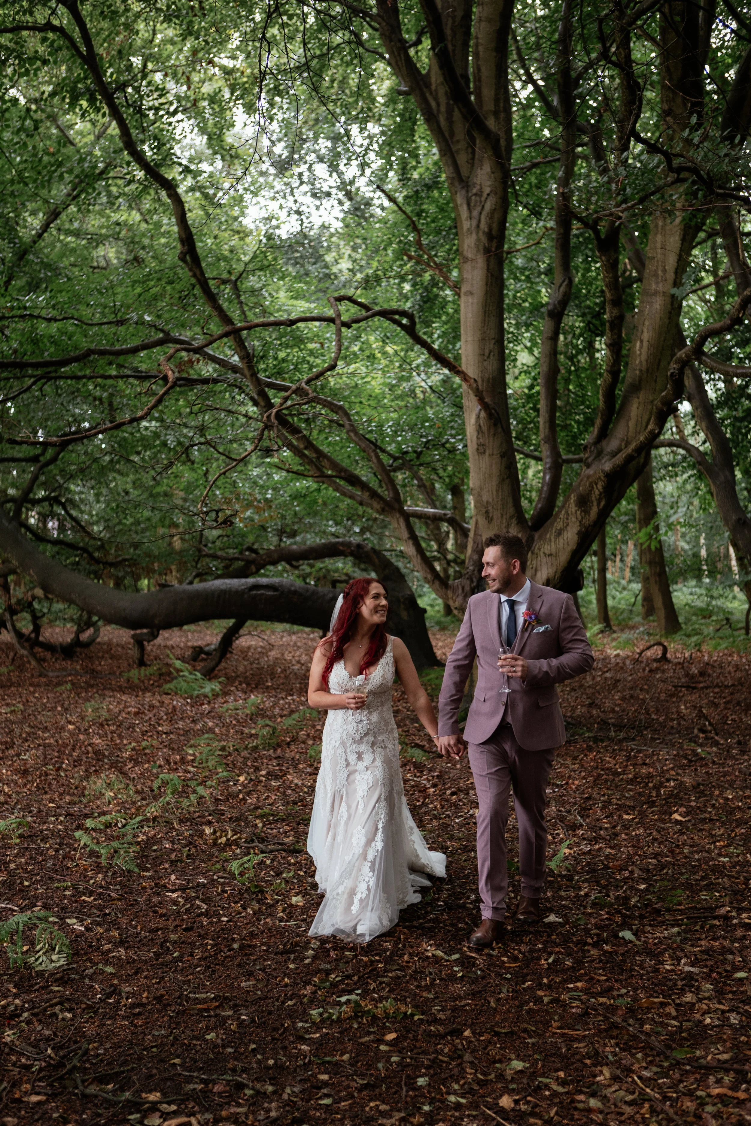 A relaxed and natural wedding moment captured by Laura Wilson Photography, showing a newly married couple walking hand in hand through a peaceful woodland setting. The bride wears a detailed lace gown and holds a glass while smiling warmly up at her 